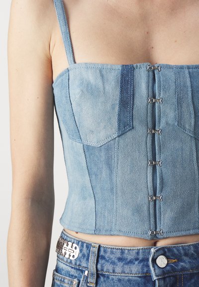 Denim crop top i lys blå farge, strukturelt design, justerbare stropper og krok-lukking foran. Brukt med blå jeans.