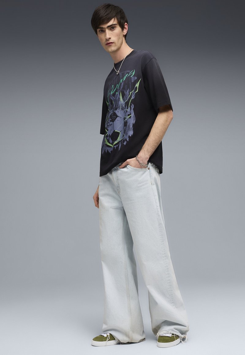 Giovane uomo che indossa una t-shirt nera con grafica, jeans chiari a gamba larga, sneakers verdi e bracciale a catena d'argento su sfondo grigio.