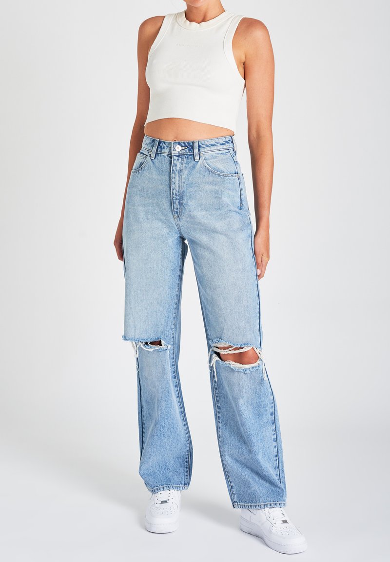 Calças de ganga de cintura alta azul-claro com rasgos desgastados, corte largo, e fecho de botão, combinadas com um top cropped branco com canelado e ténis brancos.