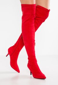 Bottes cuissardes en daim rouge avec un embout pointu et un talon stiletto. Présentent un design froissé et une fermeture éclair latérale.