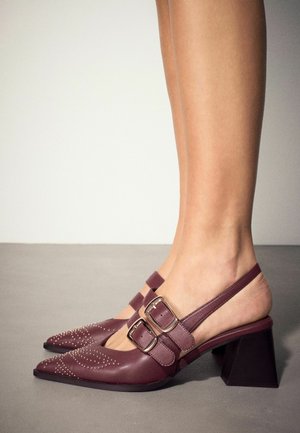 Nogi w bordowych butach na obcasie typu slingback z ozdobnymi ćwiekami, podwójnymi paskami z klamrami i grubym obcasem na szarej podłodze.