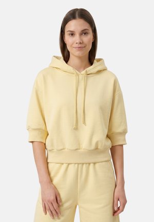 Frau mit glattem braunem Haar, die einen blassgelben Kurzarm-Hoodie und passende Hose trägt, steht vor einem schlichten hellen Hintergrund.