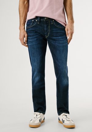 Jeans de mezclilla azul oscuro con corte recto, con un ligero desgaste, detalles en costura amarilla, cinco bolsillos y cierre de botón. Combinados con zapatillas blancas.
