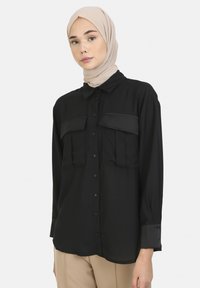 Black button-up shirt with two large chest pockets. Long sleeves with buttoned cuffs and smooth texture. Underscarf in a neutral tone.