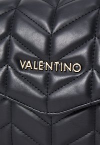 Vettävä musta nahkalaukku, jossa on shevron-malli, kullanvärinen "Valentino"-logo ja ommeltu yksityiskohta. Kompakti, teksturoitu pinta.
