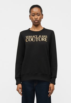 Kobieta ubrana w czarną bluzę z złotym napisem „VERSACE JEANS COUTURE”, w ciemnoniebieskich dżinsach, stojąca na jednolitym tle.