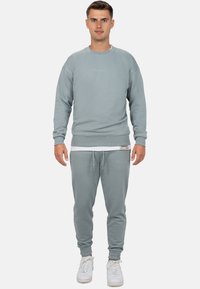 Felpa grigia chiara e pantaloni jogging abbinati, realizzati in tessuto morbido, con polsini elastici e cuciture decorative. Sneakers bianche completano il look.