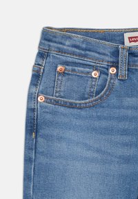 Modré džínsy z denimu s piatimi vreckami, medenými kovovými detailmi, viditeľným prešívaním a značkovou etiketou na pások.