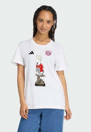 Kobieta ubrana w białą koszulkę FC Bayern Monachium z papugą trzymającą trofeum i logo klubu na jasnym tle.