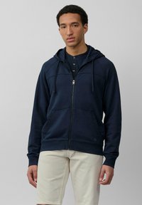 Felpa con zip di colore blu navy, dotata di cappuccio, tasche frontali e polsini a costine. Presenta una texture liscia e dettagli delle cuciture sottili. Indossata sopra una maglietta scura.