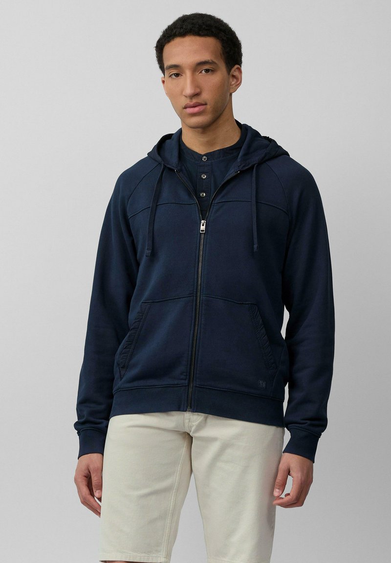 Felpa con zip di colore blu navy, dotata di cappuccio, tasche frontali e polsini a costine. Presenta una texture liscia e dettagli delle cuciture sottili. Indossata sopra una maglietta scura.