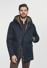 Mężczyzna z brodą ubrany w granatową kurtkę parka z kapturem, ciemną koszulę i brązowe spodnie, stojący z jedną ręką w kieszeni na białym tle.