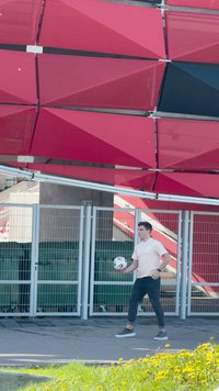 Oseba s nogometno žogo v roki stoji na pločniku ob kovinski ograji z zeleno podlago. V ozadju so rdeče in črne geometrijske okrasne plošče.