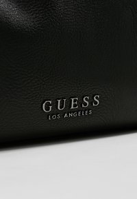 Surface noire texturée avec logo argenté en relief "GUESS LOS ANGELES" centré près du bord inférieur.