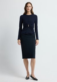 Kobieta ubrana w granatową dopasowaną bluzkę z długim rękawem, ozdobioną marszczeniami z przodu, czarną spódnicę ołówkową i czarne buty na średnim obcasie, stojąca przed białą ścianą.