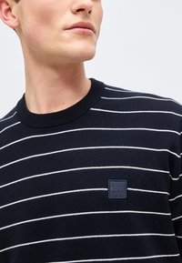 Άνδρας που φοράει μαύρο πουλόβερ με λαιμόκοψη, λεπτές λευκές οριζόντιες ρίγες και ένα μικρό τετράγωνο σχέδιο με την ένδειξη «BOSS» στο στήθος.