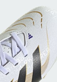 White soccer shoe featuring a textured surface, black and gold accents, and white laces. Notable logo details are visible on the side.