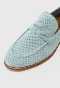 Mocasín de ante perforado azul claro con un forro de cuero suave y una suela de goma marrón. Presenta un detalle decorativo de costura en la parte superior y un diseño de fácil colocación.