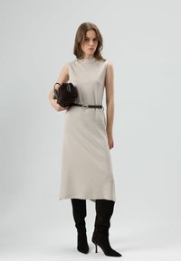 Beige geribbelde mouwloze jurk tot op de knie, gecombineerd met een donkerbruine tas en zwarte over-the-knee laarzen met hoge hakken. Bruine riem met ovale gesp.