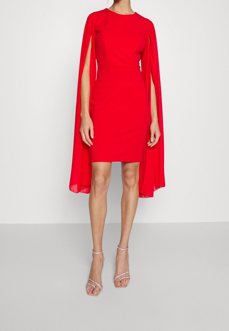 Femme portant une robe rouge vif ajustée longueur genou avec manches cape et sandales à talons argentées sur un fond uni.