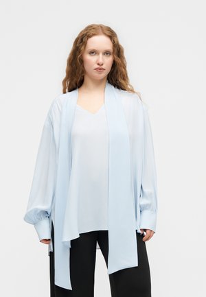 Mlada ženska s kodrastimi rdečimi lasmi, oblečena v ohlapno svetlo modro bluzo in črne hlače, stoji pred enobarvnim belim ozadjem.