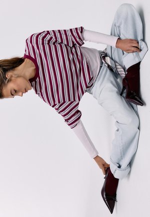 Frau sitzt auf dem Boden, trägt ein bordeauxrotes und weiß gestreiftes Shirt, helle Jeans und dunkle spitze Schuhe und richtet mit der Hand einen Schuh.