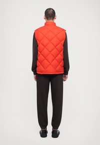 Rød quiltet puffervest med diamantmønster og høj krave, båret over et mørkt outfit, med en afslappet pasform og sidesøm.