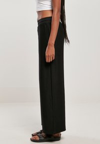 Pantalon large en côte noir avec une taille élastique, associé à des sandales plates noires. Le tissu a une finition texturée, créant un intérêt visuel.