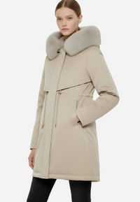 Parka beige con colletto in pelliccia sintetica, chiusure con zip e cordoncino, tasche laterali e design elegante. Materiale liscio con finitura opaca.