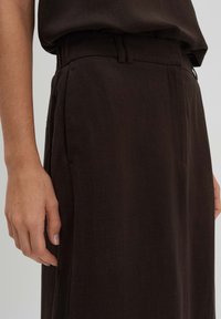 Pantalons en lin marron avec une taille haute et une coupe slim, dotés de poches latérales et d'une texture subtile. Main placée sur le côté pour référence.