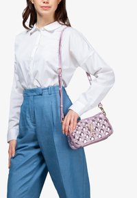 Bolso de mano metálico rosa con patrón de burbujas texturizado, correa ajustable y detalle de logo. Usado con una camisa blanca y pantalones azules.