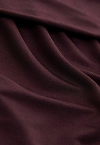 Burgundijas stiepļu audums ar gludu, teksturētu virsmu, kurā redzami smalki ribojumi. Materiāls dabiski krīt ar elegantiem krokām.