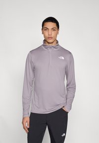 Πουλόβερ με μακριά μανίκια σε ανοιχτό μωβ ύφασμα με φερμουάρ στο λαιμό, με το λογότυπο The North Face σε λευκό στην αριστερή πάνω πλευρά του στήθους.