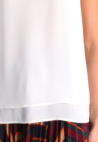 Blouse blanche au tissu lisse, avec un ourlet superposé. Elle est associée à une jupe à motifs aux couleurs vives avec des formes géométriques.