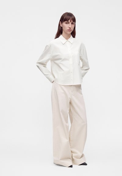 Femme debout, les mains derrière le dos, portant une chemise blanche à boutons, un pantalon beige large et des chaussures noires pointues sur fond blanc.