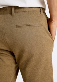 Pantaloni marroni, con texture e slim fit, con tasca posteriore e passanti per cintura, mostrati dalla vita in giù.