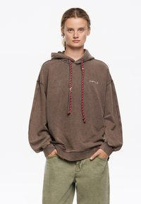 Sudadera oversized marrón con un logo frontal, que presenta cordones en rojo y negro. Combinada con pantalones verde claro, que muestran un ajuste relajado.