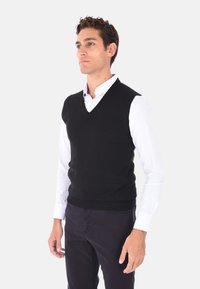 Gilet nero in maglia con scollatura a V, indossato sopra una camicia bianca con colletto. Texture liscia e dettagli a coste sull'orlo.