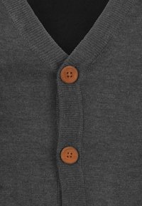 Grå strikket cardigan med V-hals, med to brune knapper. Teksturen ser myk ut, og stoffet har en glatt overflate.