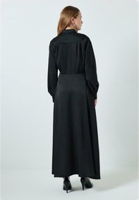 Black maxi dress with long sleeves, a buttoned collar, and a tailored waist. Smooth, matte texture with minimalist design. Heels visible.