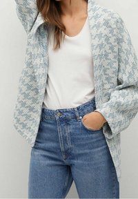 Jasnoniebieska kurtka w wzory z teksturowanej tkaniny, z otwartym przodem i luźnymi rękawami. Noszona na białej bluzce i zestawiona z dżinsami z wysokim stanem.