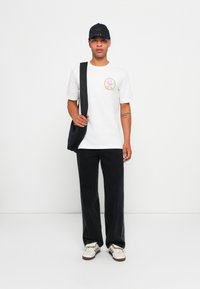 T-shirt branca de algodão com um gráfico redondo colorido, combinada com calças largas pretas e um boné preto. O modelo segura uma mala de ombro preta.