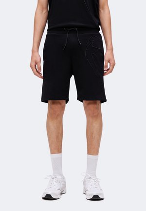 Homme portant un short noir au genou avec cordon, des chaussures de sport blanches et des chaussettes blanches, debout devant un fond uni.
