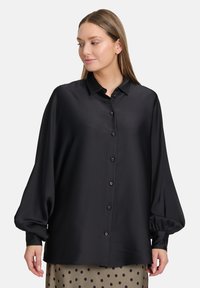 Camicia nera in satin con colletto, maniche a palloncino oversize e taglio dritto, abbinata a una gonna a pois.
