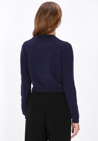 Granatowy sweter z ribbowymi mankietami i skróconym krojem. Posiada fakturowane rękawy w splot kablowy, ozdobione małymi, dekoracyjnymi koralikami.
