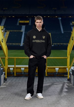 Jonge man in een zwarte Guess Jeans hoodie en broek staat met zijn handen in zijn zakken in een leeg stadion met gele zitplaatsen.