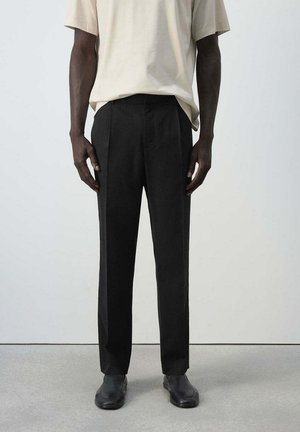 Hombre con pantalones negros a medida, zapatos negros y una camisa beige de manga corta, de pie sobre un suelo gris claro frente a una pared blanca lisa.