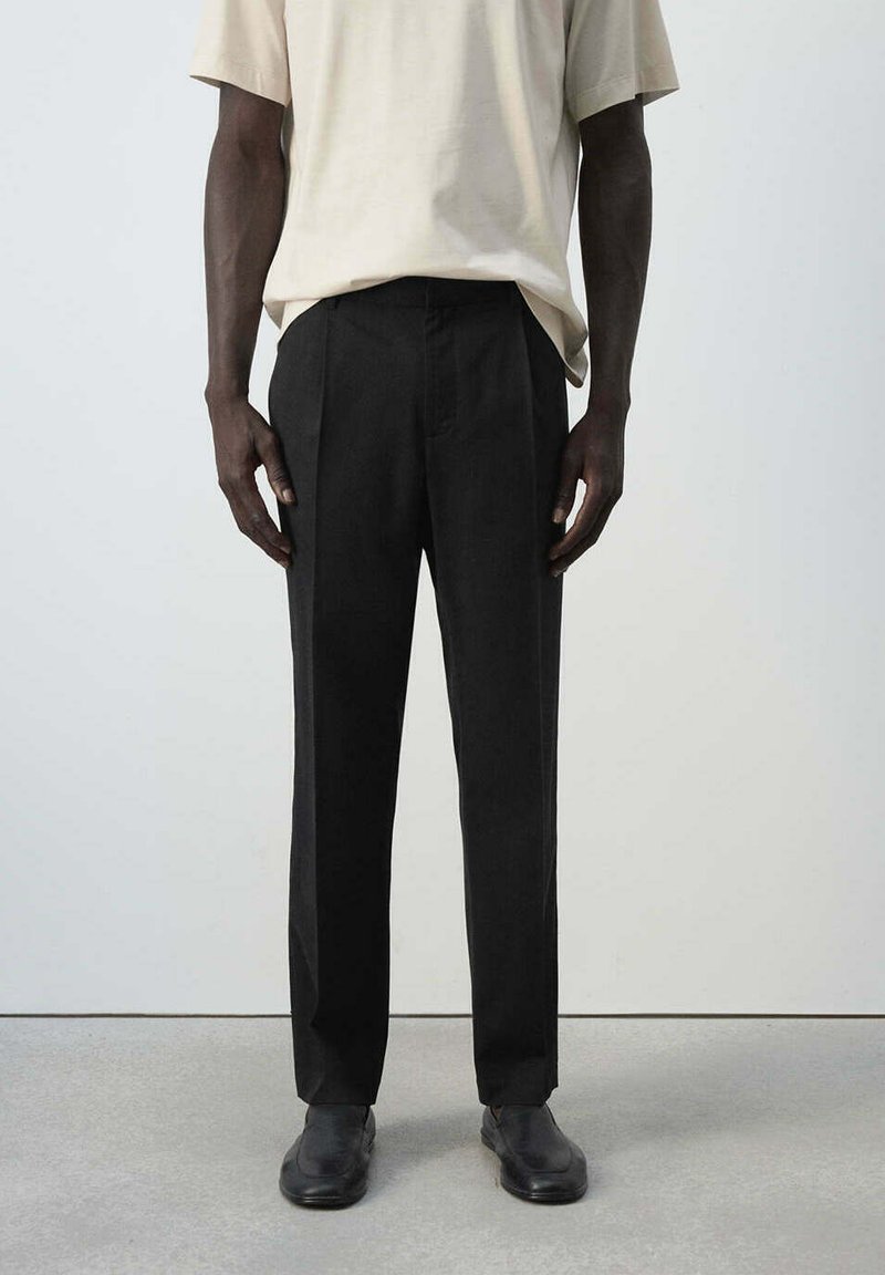 Homme portant un pantalon noir sur mesure, des chaussures noires et une chemise beige à manches courtes, debout sur un sol gris clair contre un mur blanc uni.