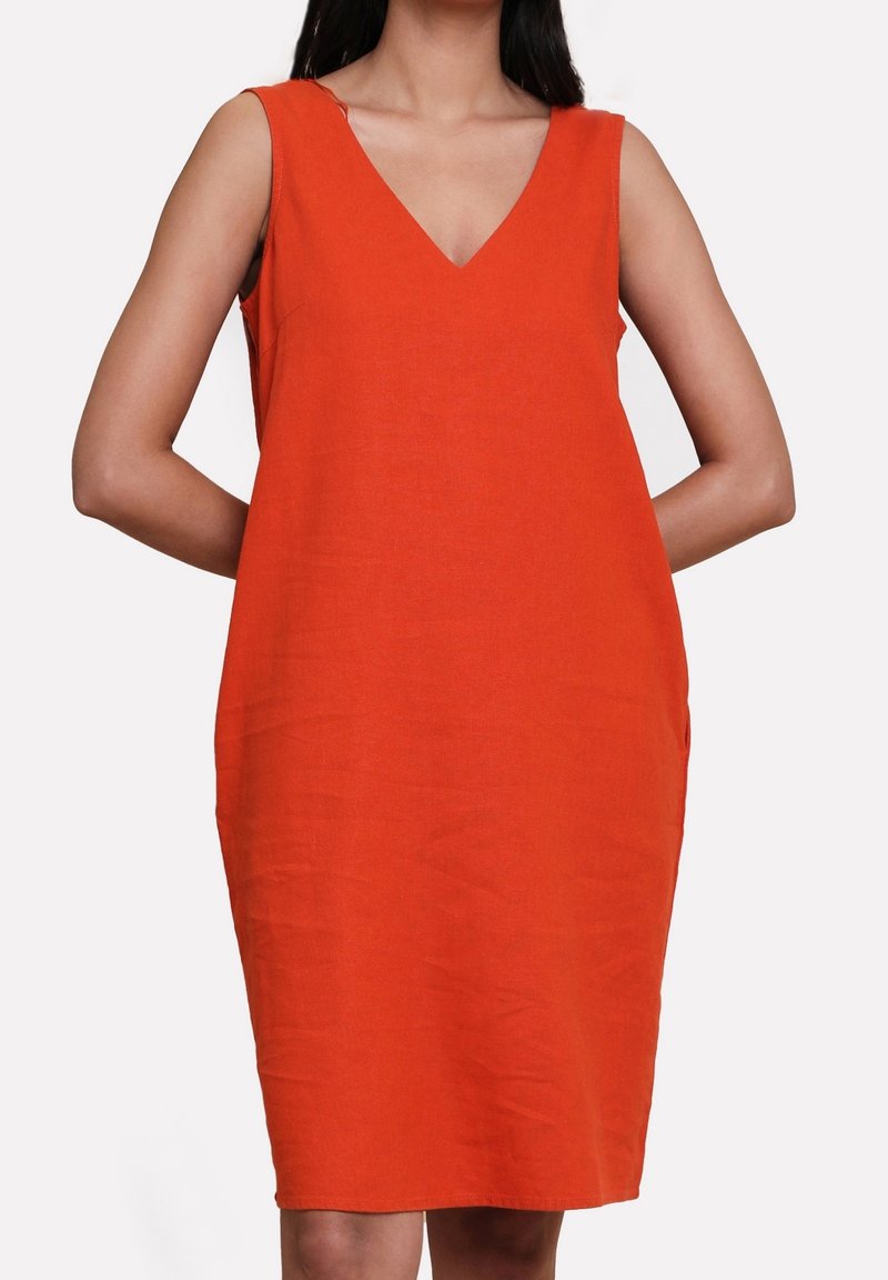 Femme portant une robe orange vif sans manches, longueur au genou, col en V, mains dans le dos sur fond blanc uni.