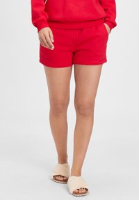 Red cotton shorts with an elastic waistband and side pockets, featuring a smooth texture and a casual design. Worn with beige fuzzy slippers.
